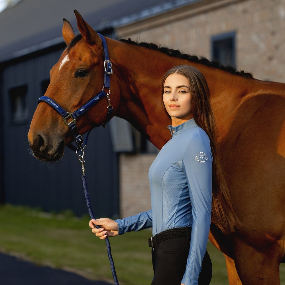 Woman in equestrian attire standing next to a brown horse in an outdoor setting.